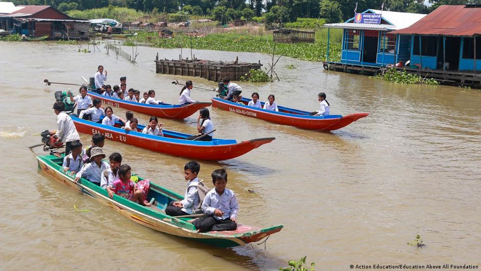 Students travel by boat past floating homes and vegetation on a broad muddy river.