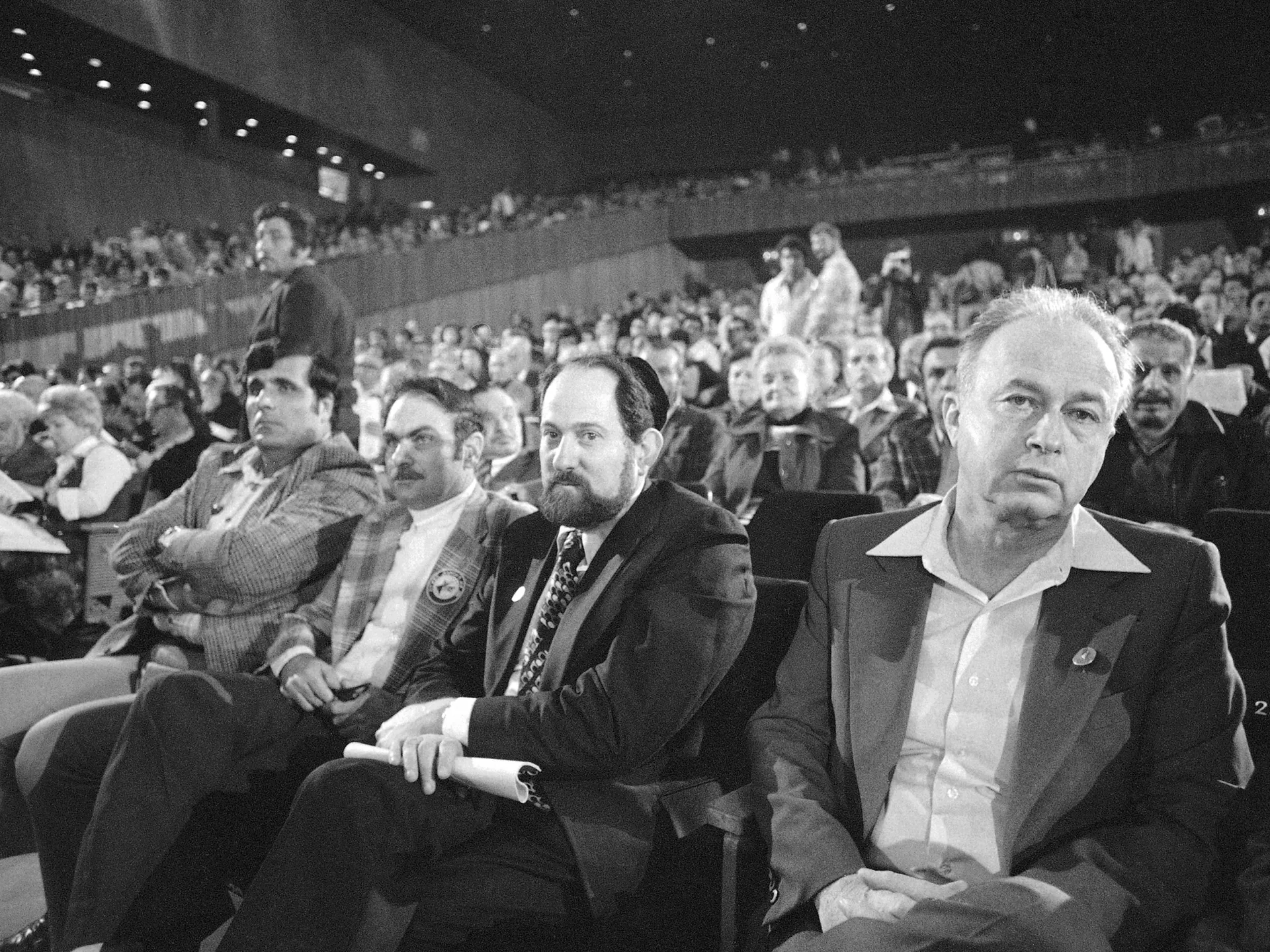 Black-and-white photograph of four men seated among a large audience at a formal event.