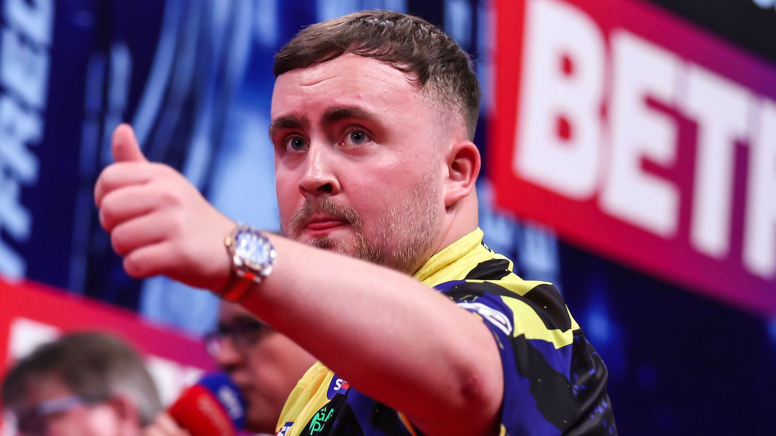 A bearded man in a yellow and black sports shirt points toward the camera, with a blurred event logo behind him.
