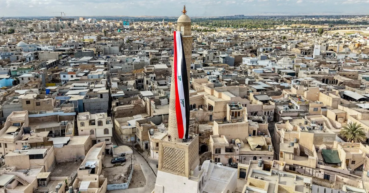 A large Iraqi flag hangs from a tall tower above a dense expanse of low-rise buildings.
