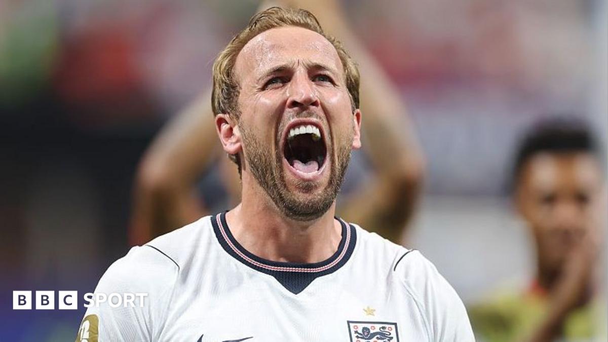 Footballer in white England shirt shouting during a match, with blurred spectators behind him.
