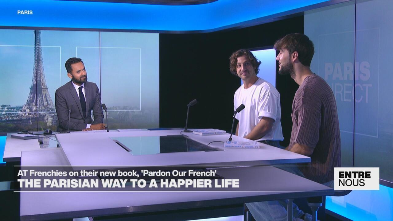 A suited presenter speaks with two male guests seated at a studio desk beneath blue Paris-themed screens.