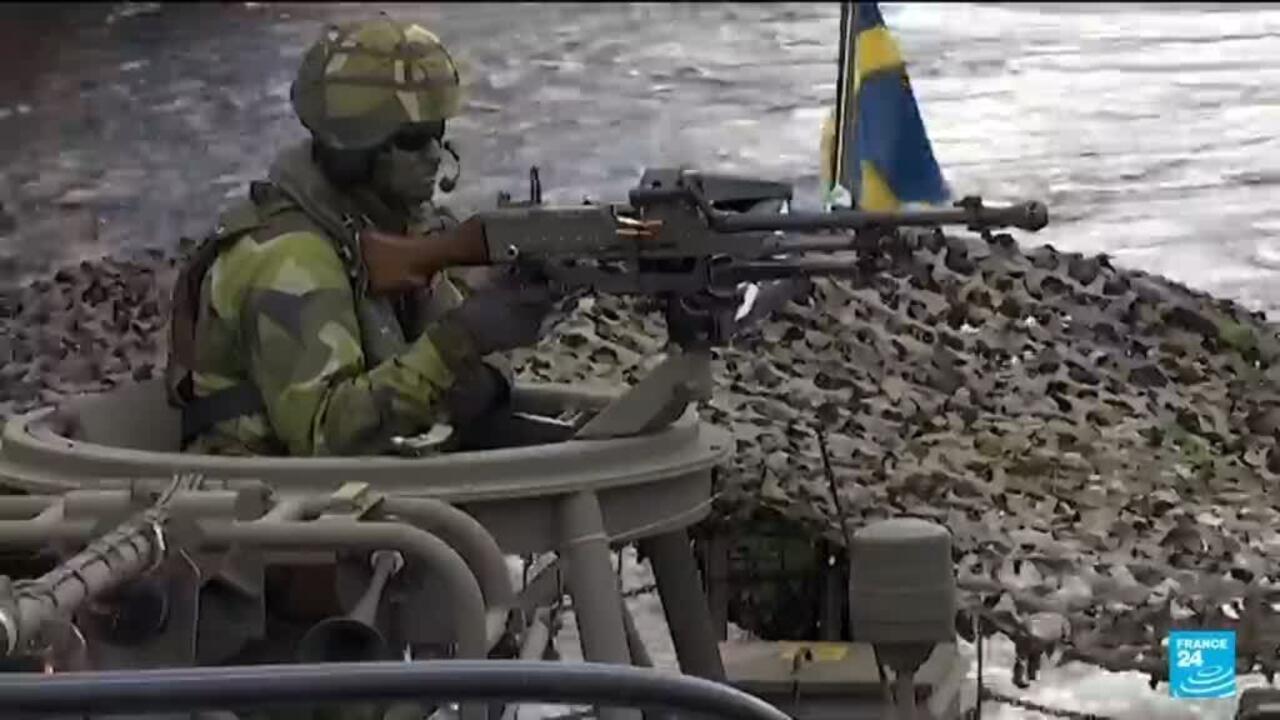 Soldier in camouflage aims a rifle from a military vehicle beside a Swedish flag