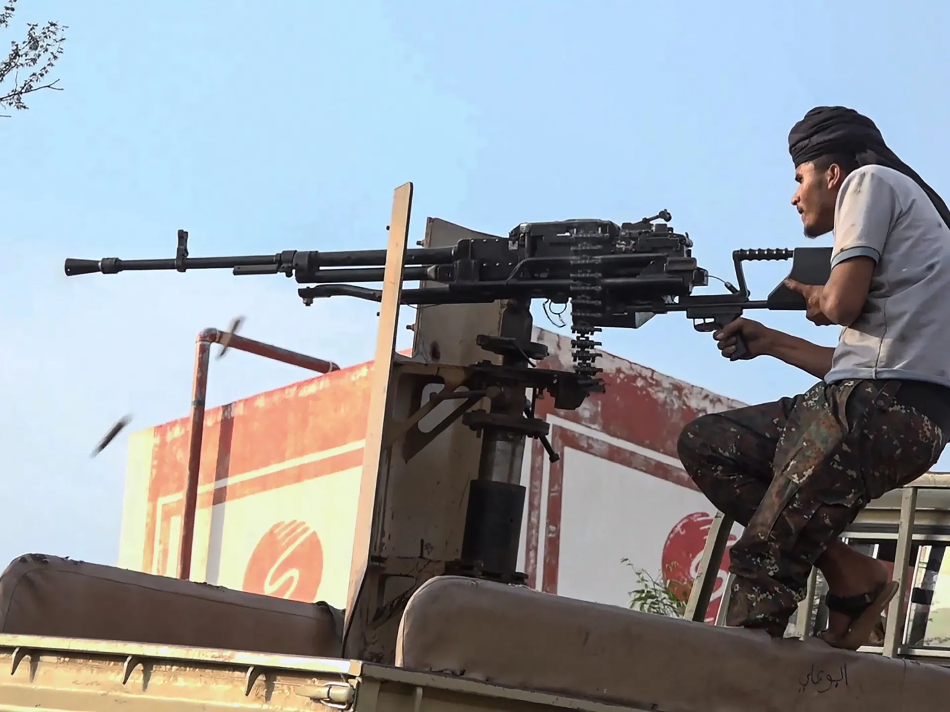 Armed man operates a mounted heavy machine gun from a vehicle under a clear blue sky.