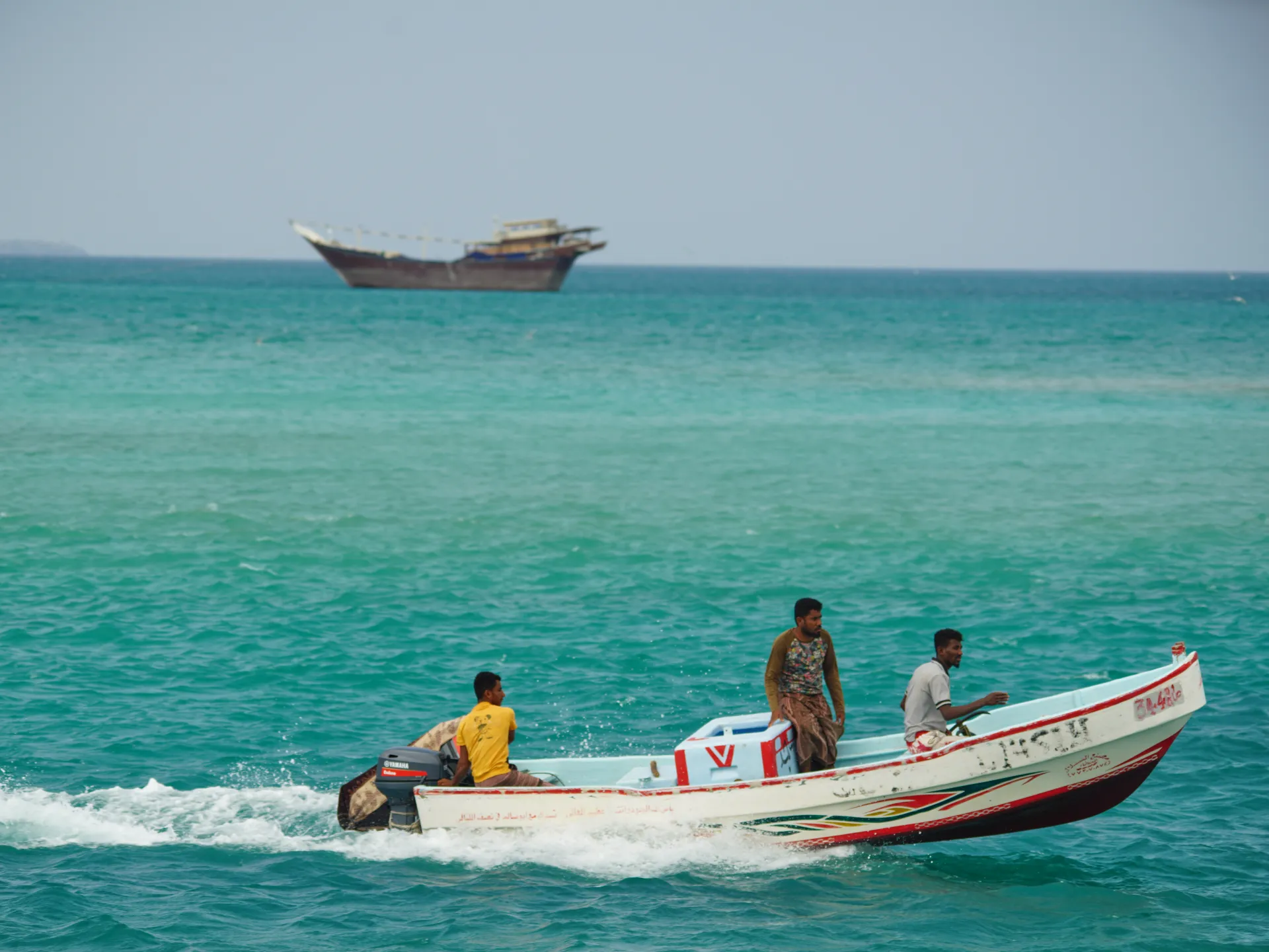 Three men travel in a small motorboat across turquoise sea waters, with another boat on the horizon.