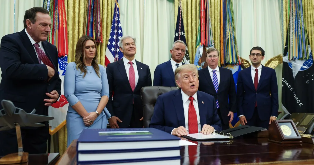 Donald Trump sits at a desk signing a document as officials stand behind him.