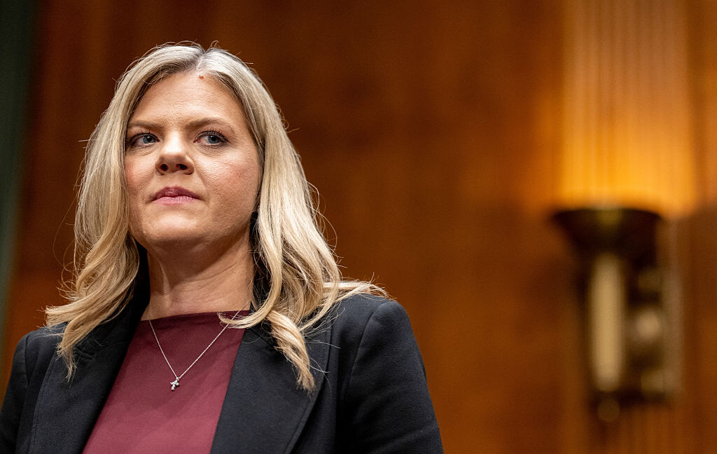 Blonde woman in a dark blazer and burgundy top stands before a warmly lit wood-paneled background.