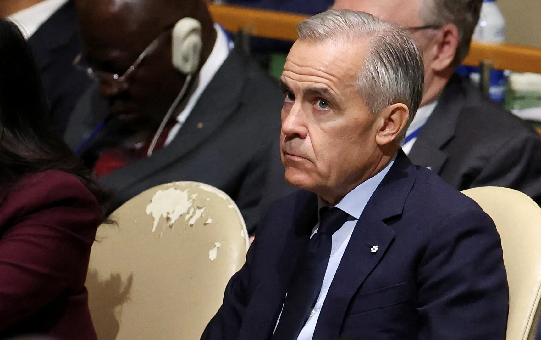 A silver-haired man in a dark suit sits among attendees at an indoor event.