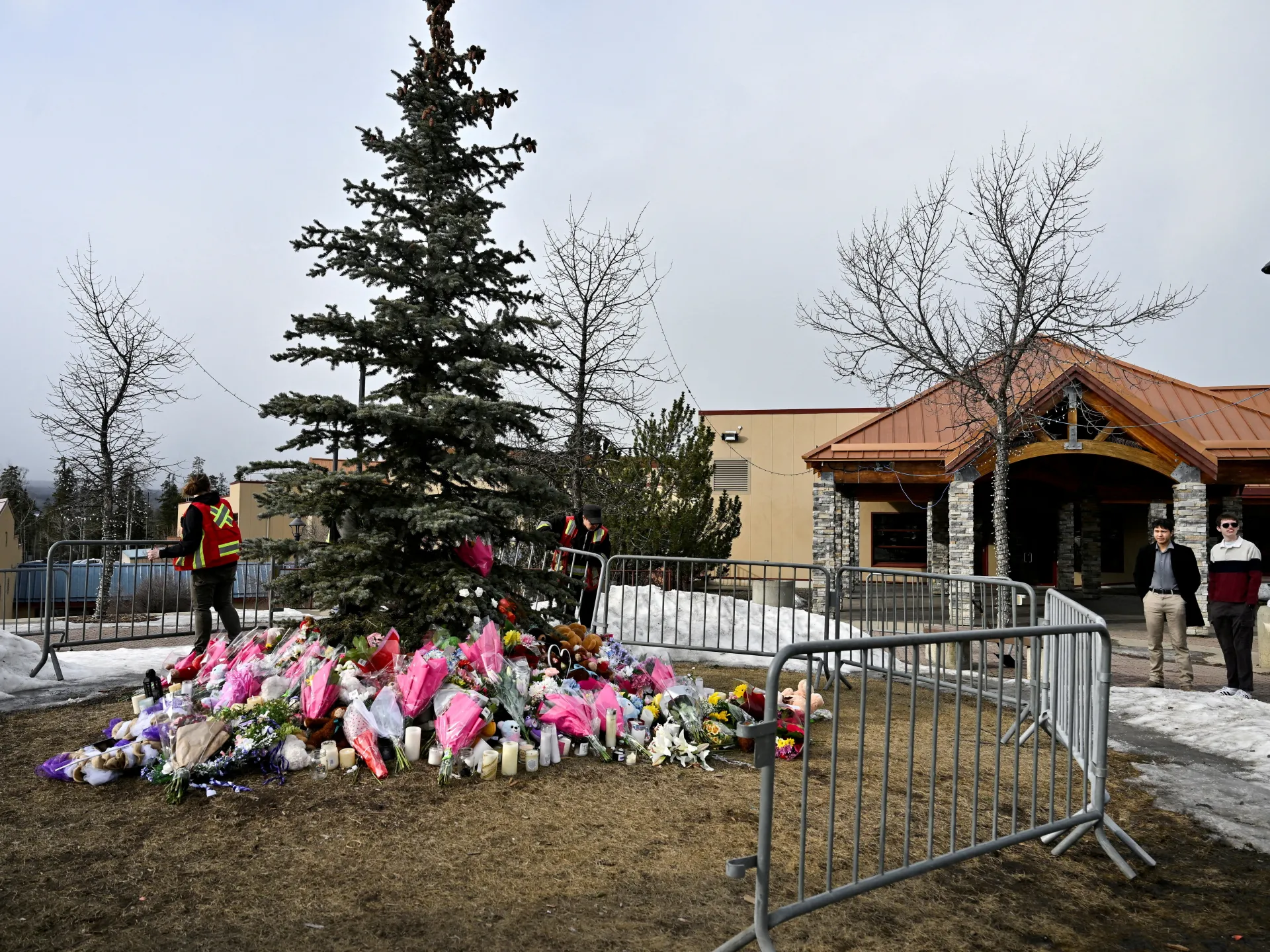 Flowers and candles surround a large evergreen memorial outside a building in snowy conditions.