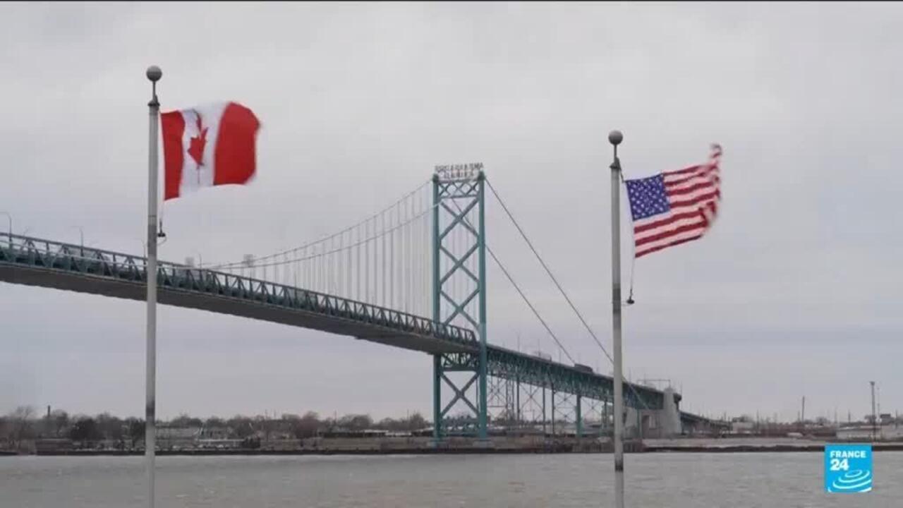 Canadian and U.S. flags fly beside a suspension bridge over a river under gray skies.