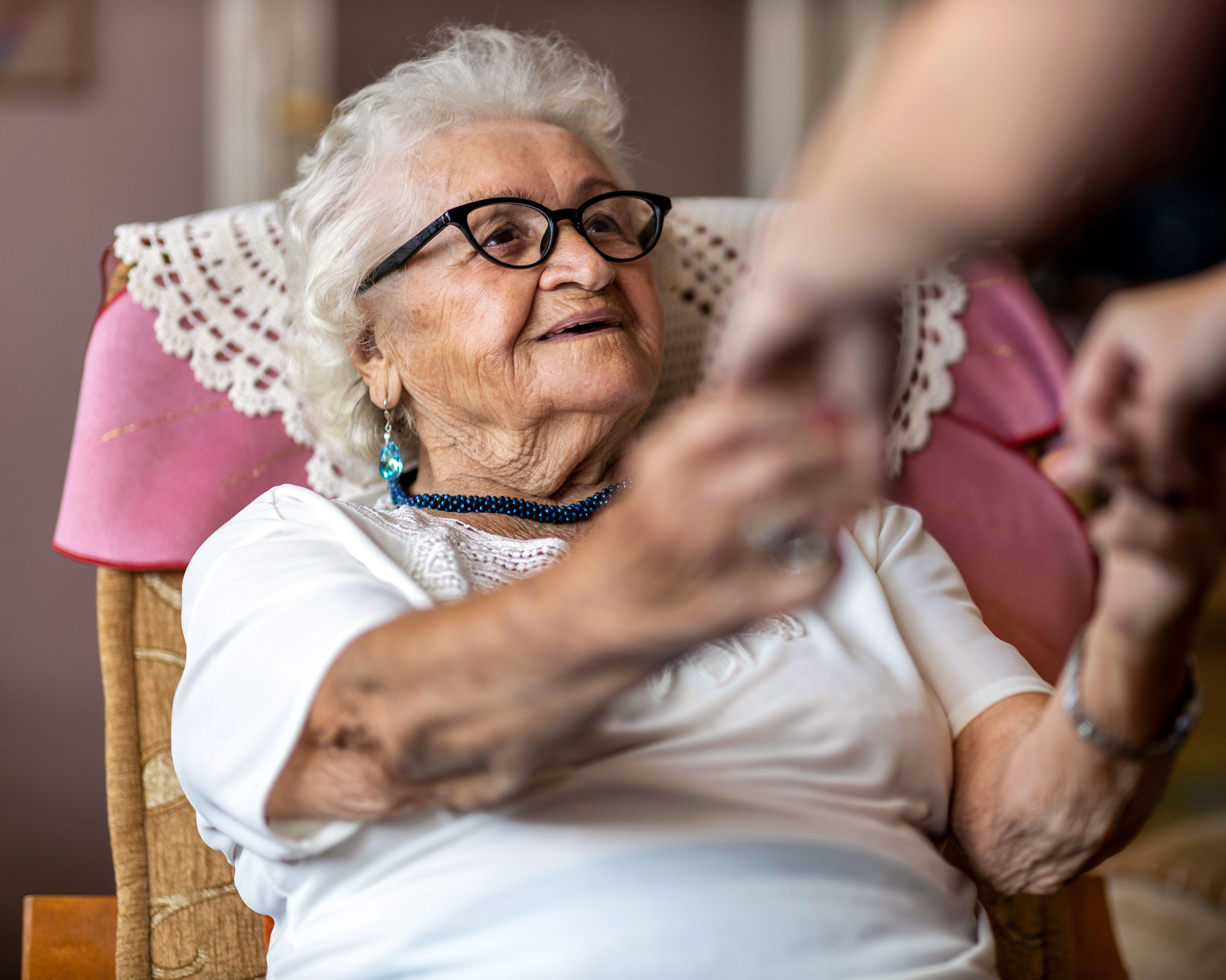Elderly woman wearing glasses sits in a chair as a blurred person reaches toward her.