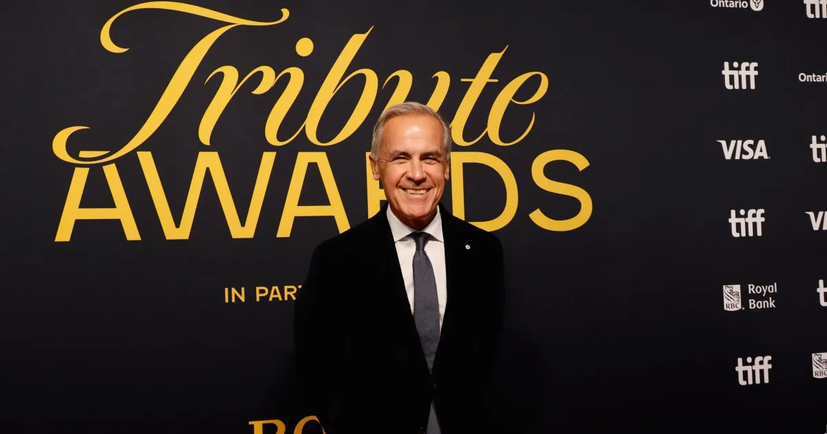 Smiling suited man stands before a black-and-gold Tribute Awards backdrop at a red-carpet event.