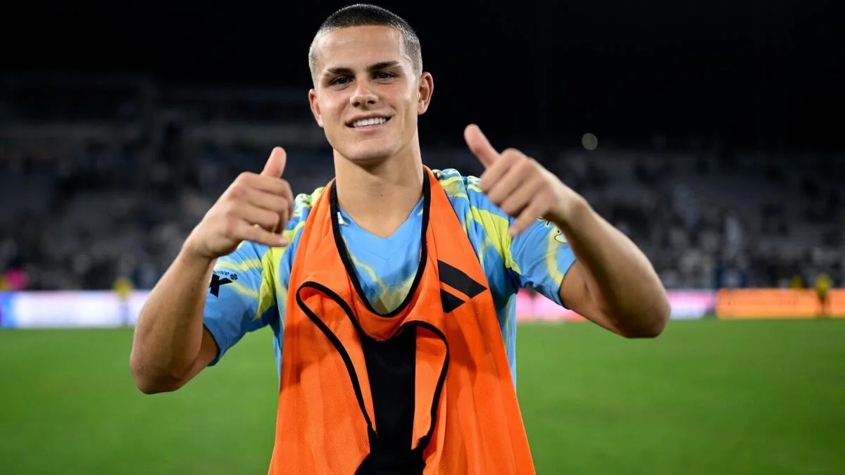 Young soccer player in an orange training bib smiles and gives two thumbs up on the field.