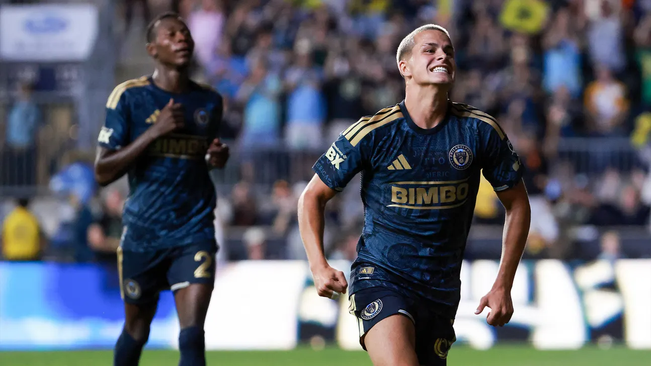 Soccer player in navy and gold jersey celebrates while running, with a teammate behind him.