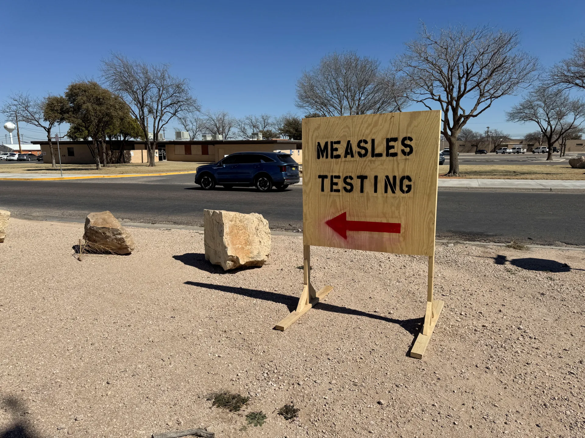 A roadside wooden sign reads “MEASLES TESTING” above a red arrow pointing left.