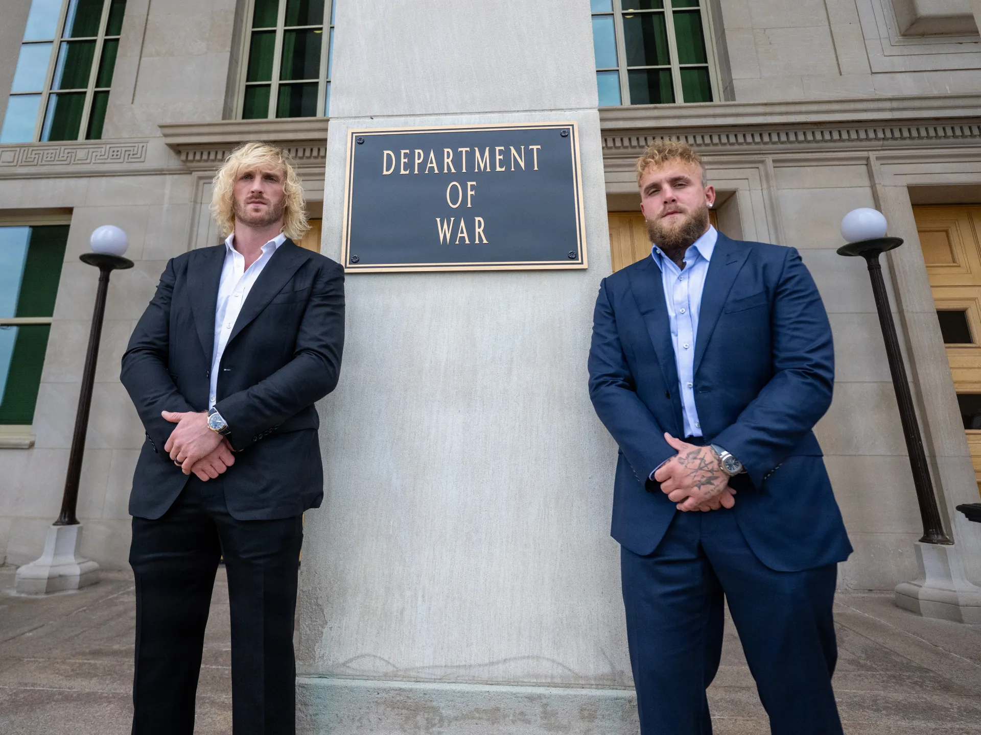 Two men in dark suits stand beside a plaque reading “Department of War.”