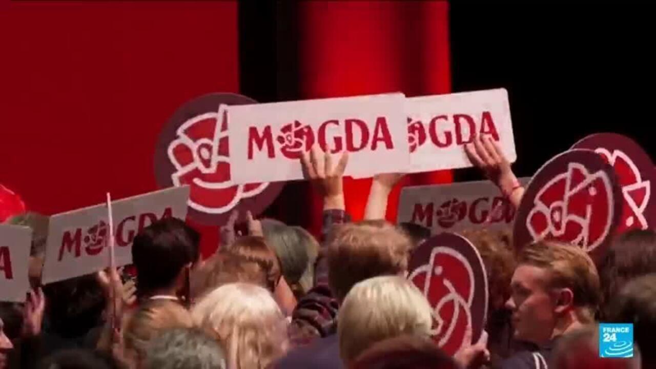 Crowd holds red-and-white political signs and symbols at a televised indoor gathering.