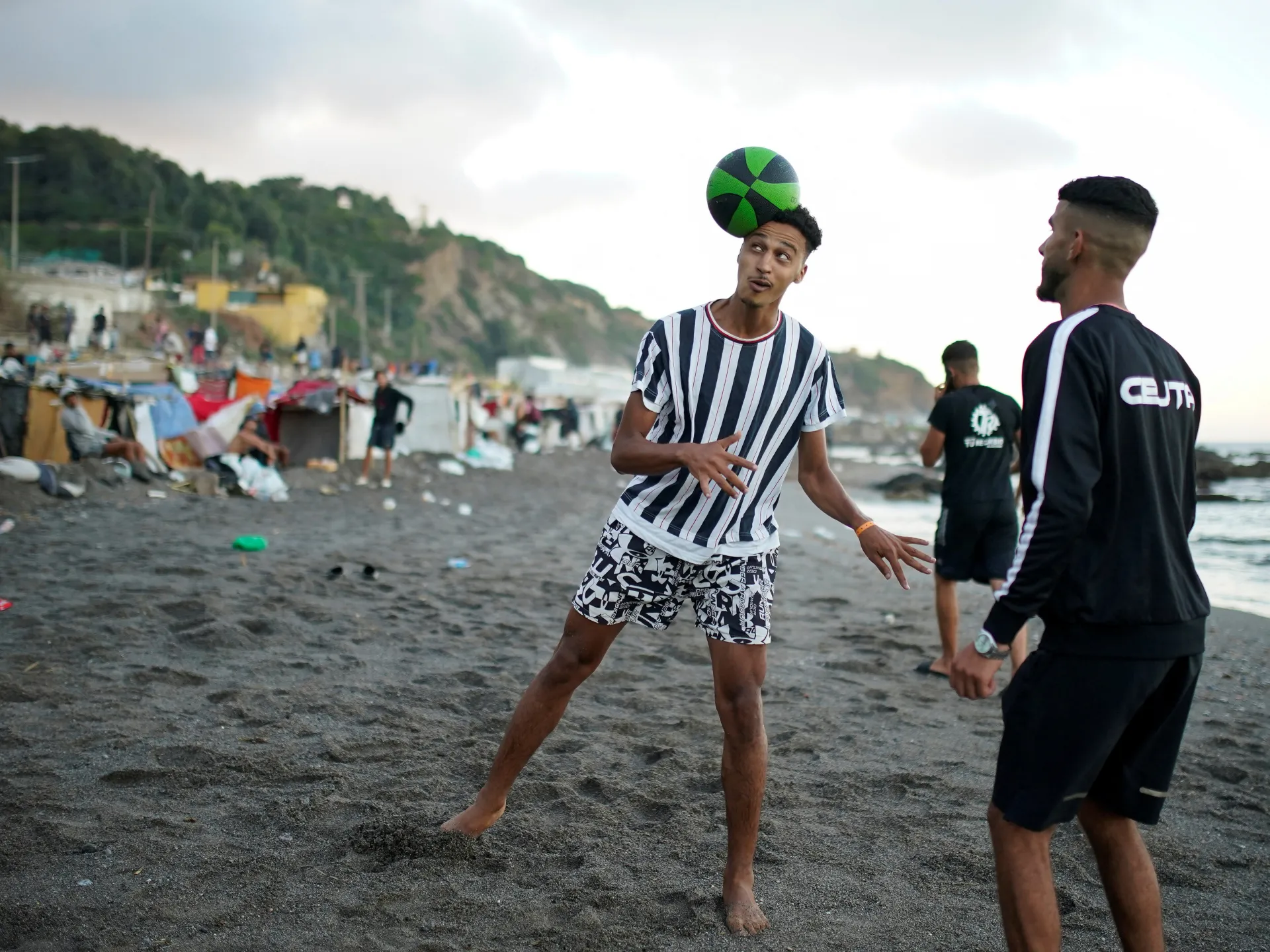 Young men play football on a crowded beach, with one balancing a green-and-black ball on his head.