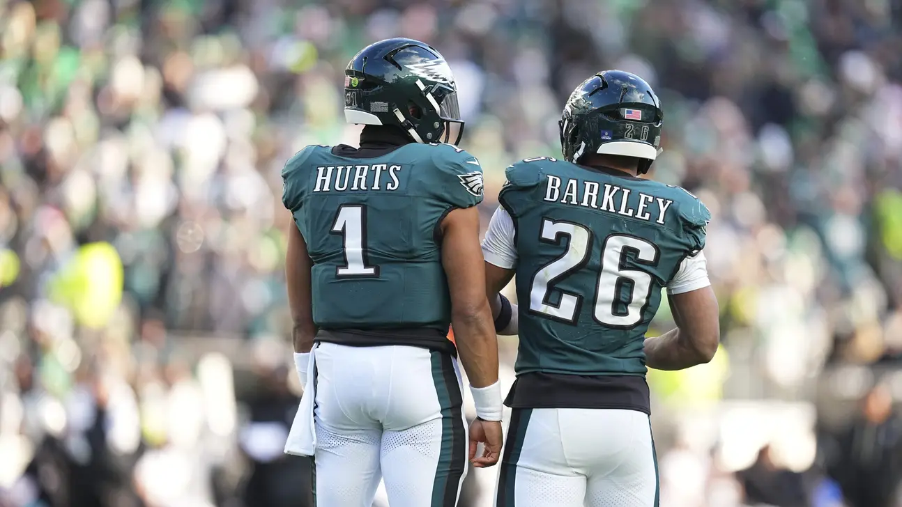 Two Philadelphia Eagles players, wearing helmets and jerseys labeled Hurts and Barkley, walk onto the field.