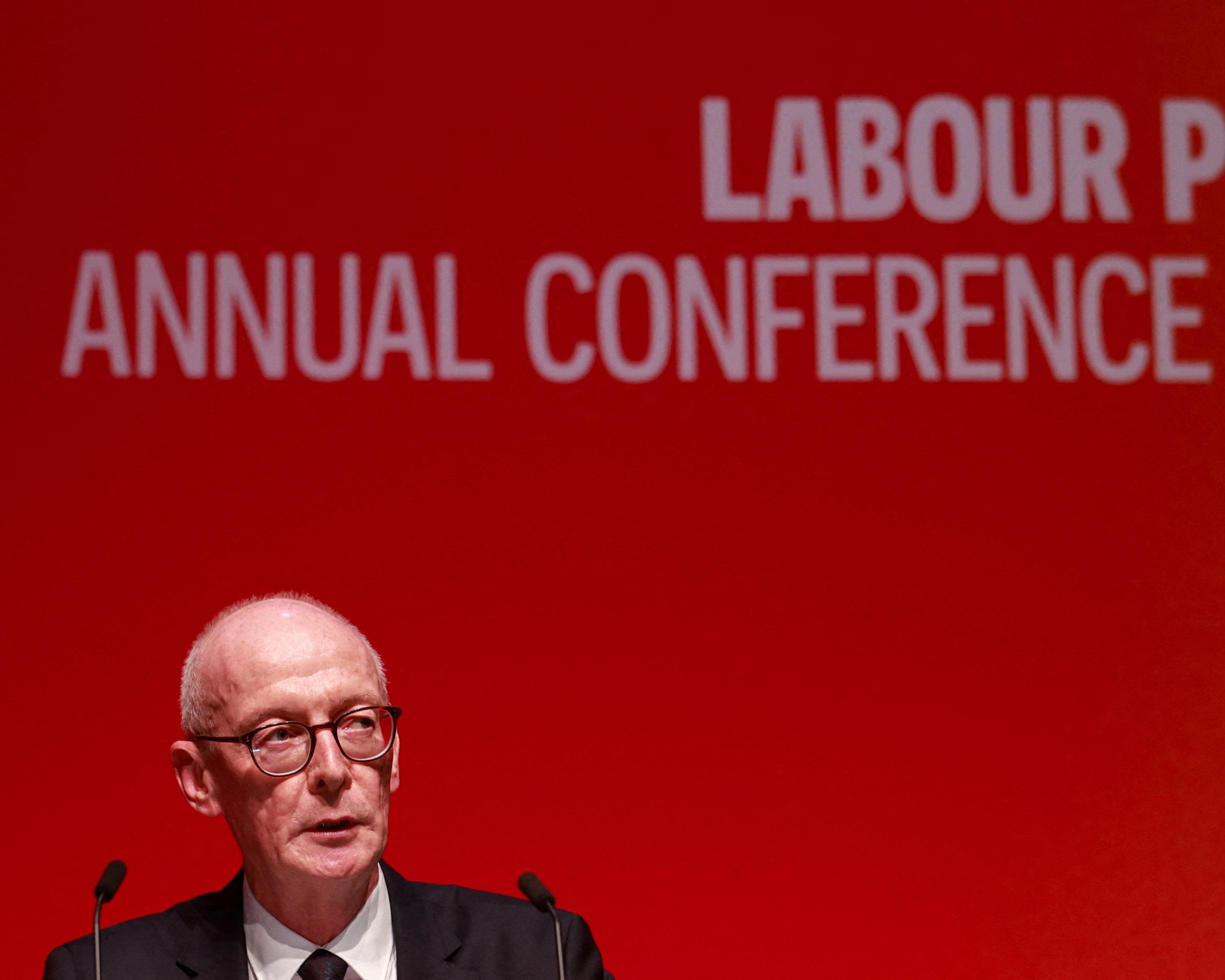 A bespectacled man speaks at microphones against a red backdrop with partial Labour conference lettering.