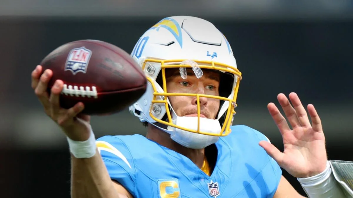 Illustrated football player in blue uniform raises a hand while holding a helmet beside his face.