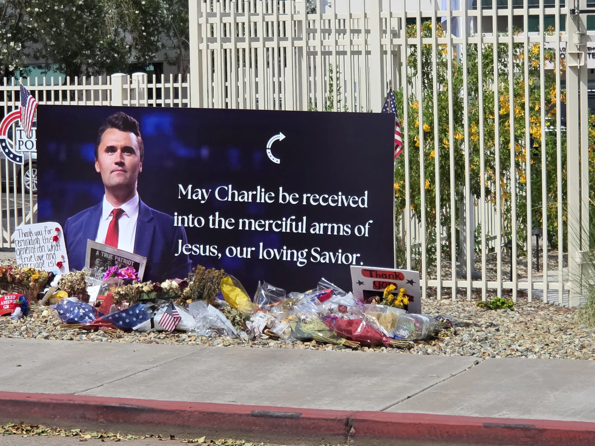 Memorial display with flowers, flags and a portrait sign outside a gated building.