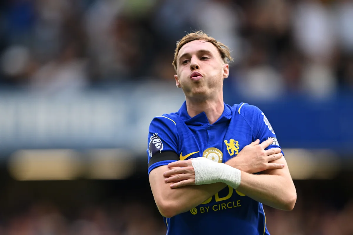 Chelsea player in blue shirt crosses his arms during a match, with stadium crowd blurred behind.