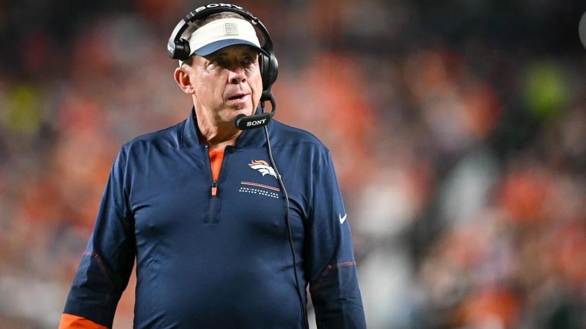 Football coach wearing a headset and navy Broncos pullover on the sideline.