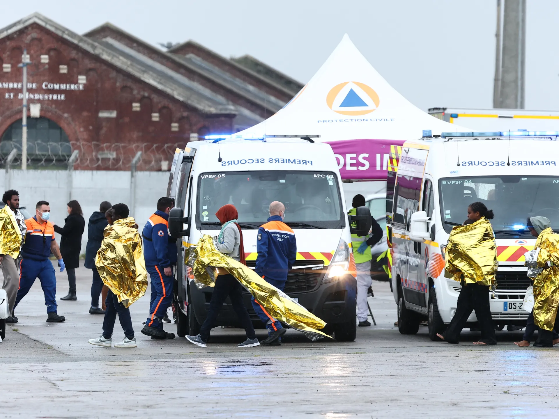 French civil protection workers assist people wrapped in gold thermal blankets beside ambulances outside a brick building.