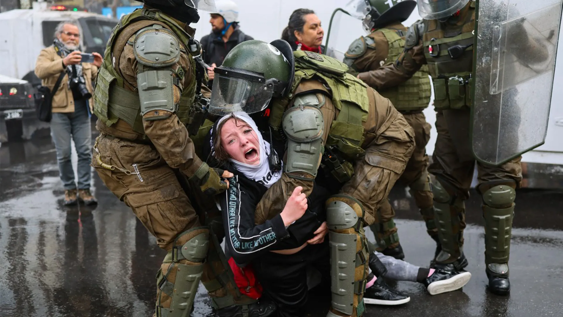 Armored police officers detain a distressed woman on a wet street as bystanders watch.