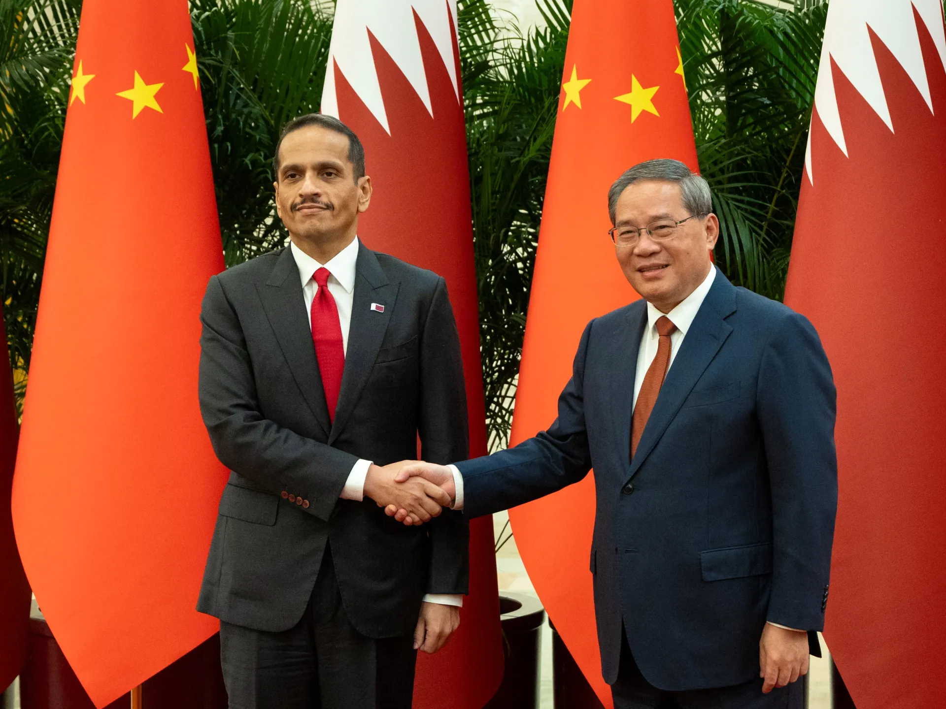 Two suited officials shake hands in front of Chinese and Qatari flags and greenery.