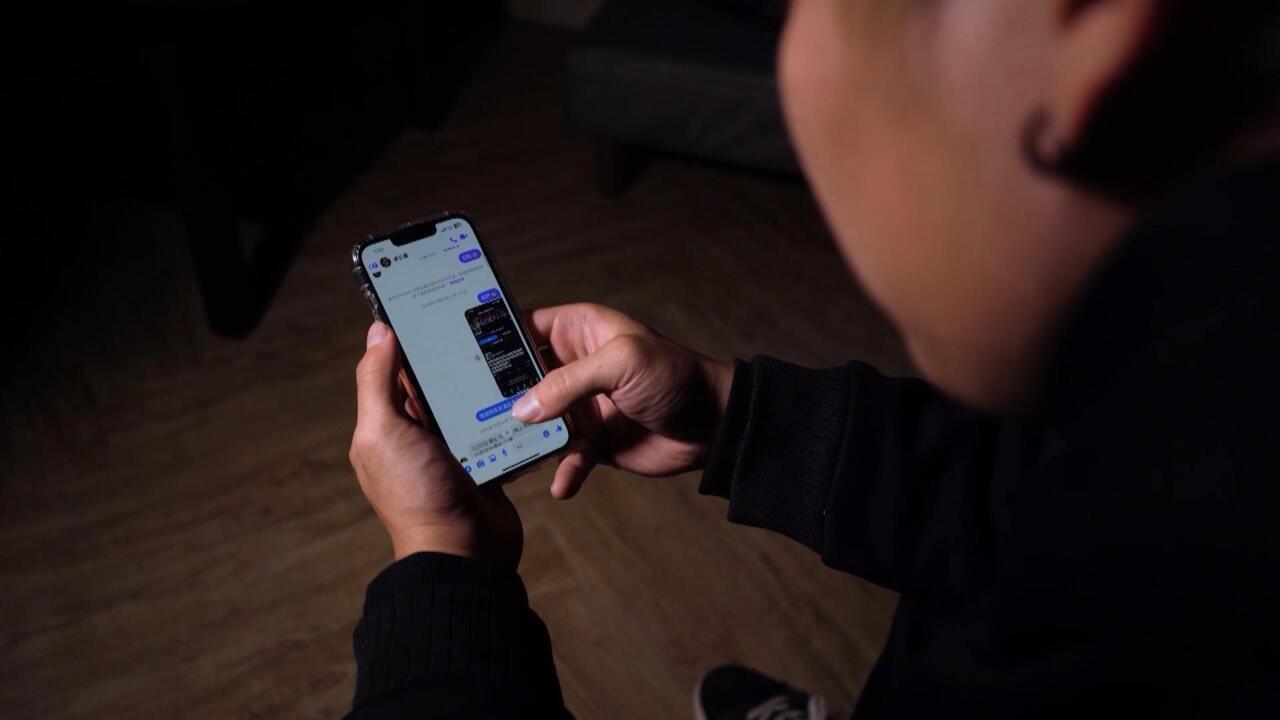 A person in dark clothing holds a smartphone and taps its screen in a dim room.