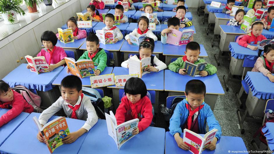 Chinese schoolchildren sit at blue desks reading books in a crowded classroom.
