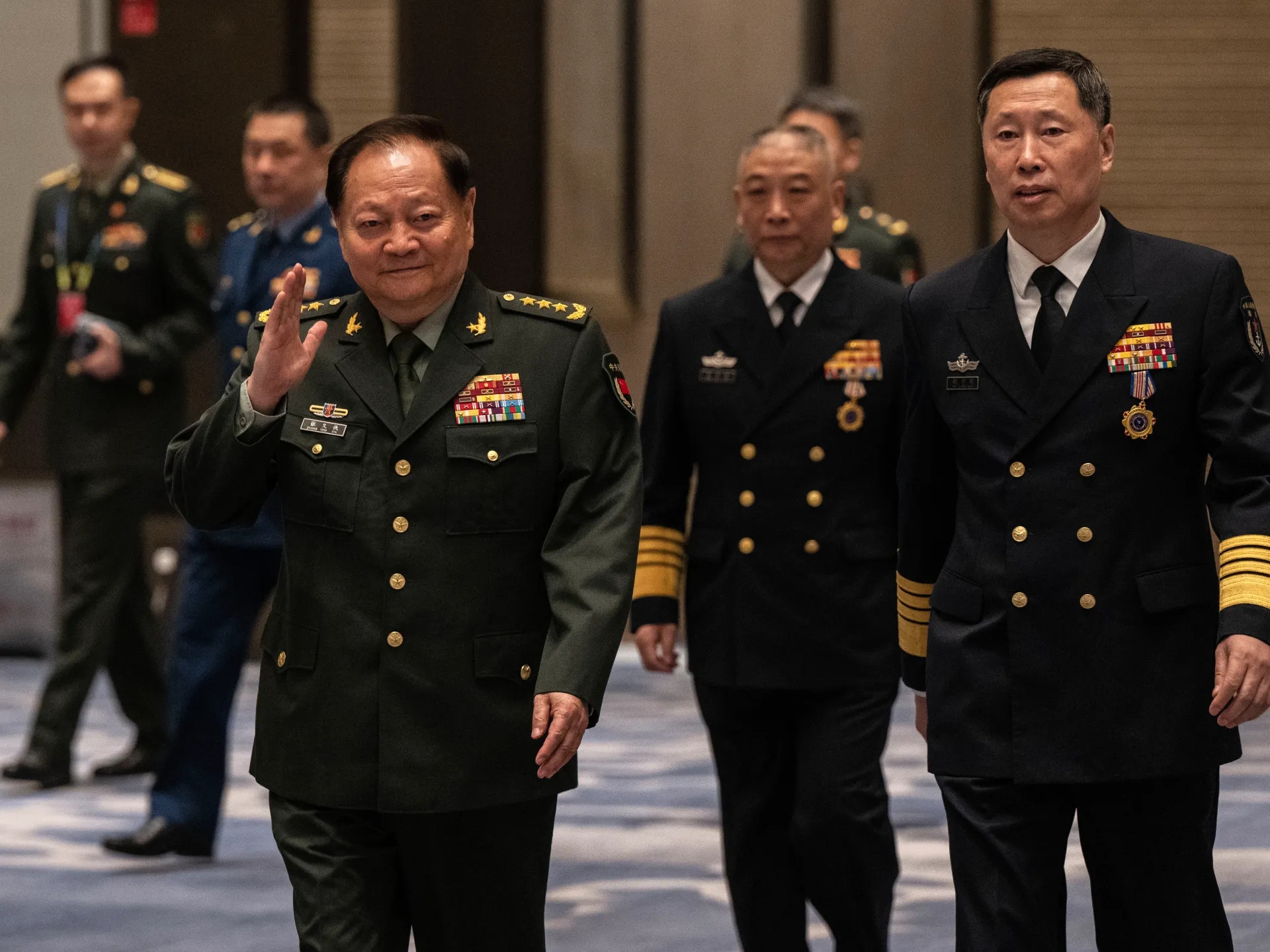 Uniformed Chinese military officials walk indoors, with one officer raising his hand in salute.