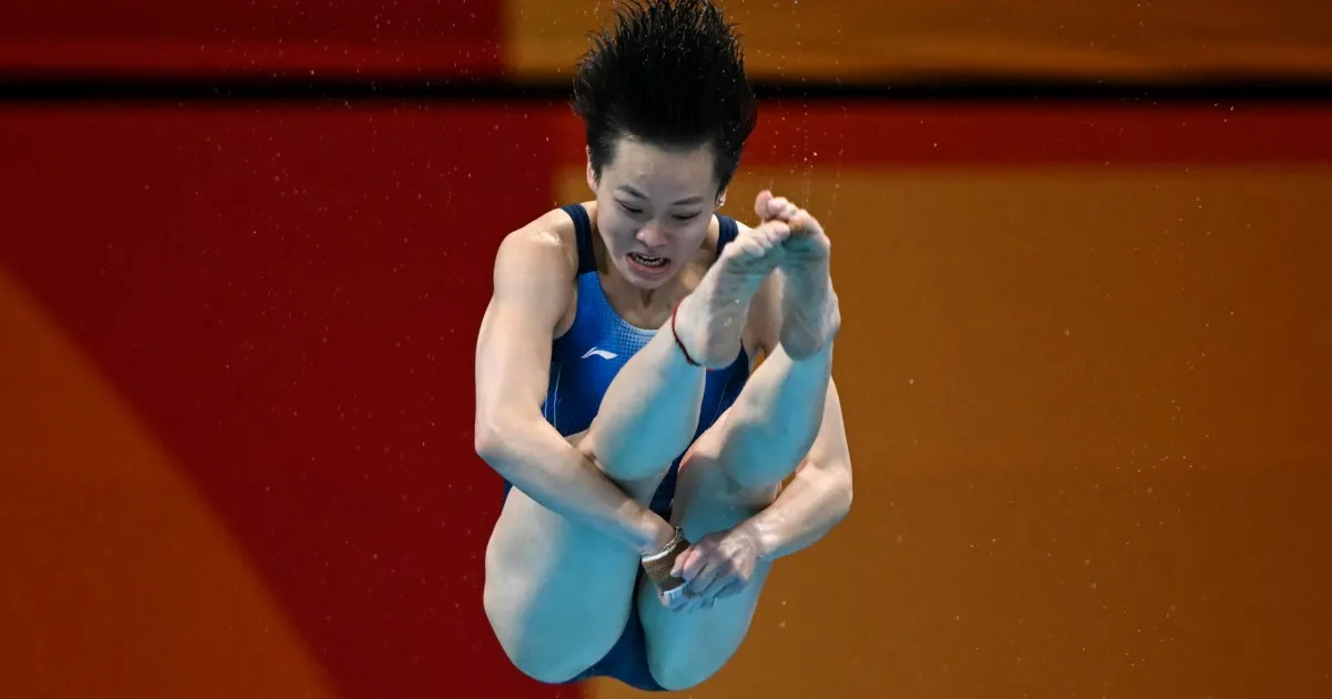 A diver in a blue suit tucks her legs during a dive, with droplets around her.