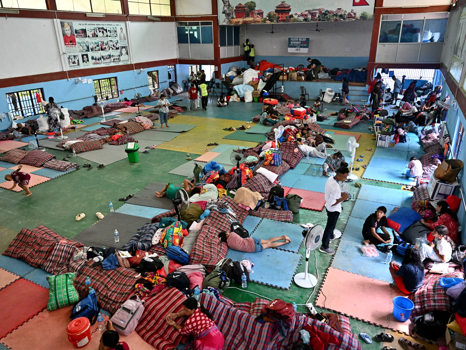Crowded flood relief shelter filled with people, bedding, belongings, fans and colorful floor mats