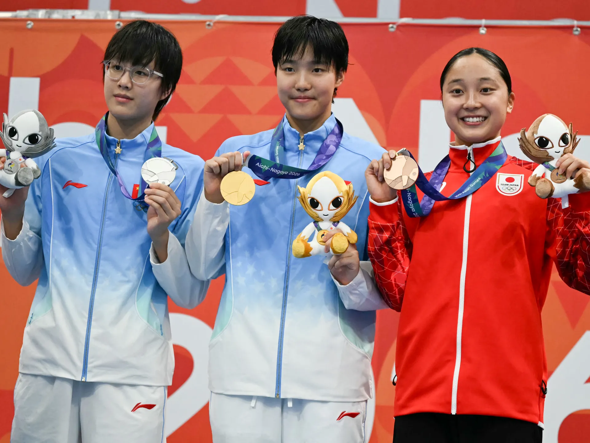 Three people in blue and red sportswear display medals and plush mascots against a red backdrop.