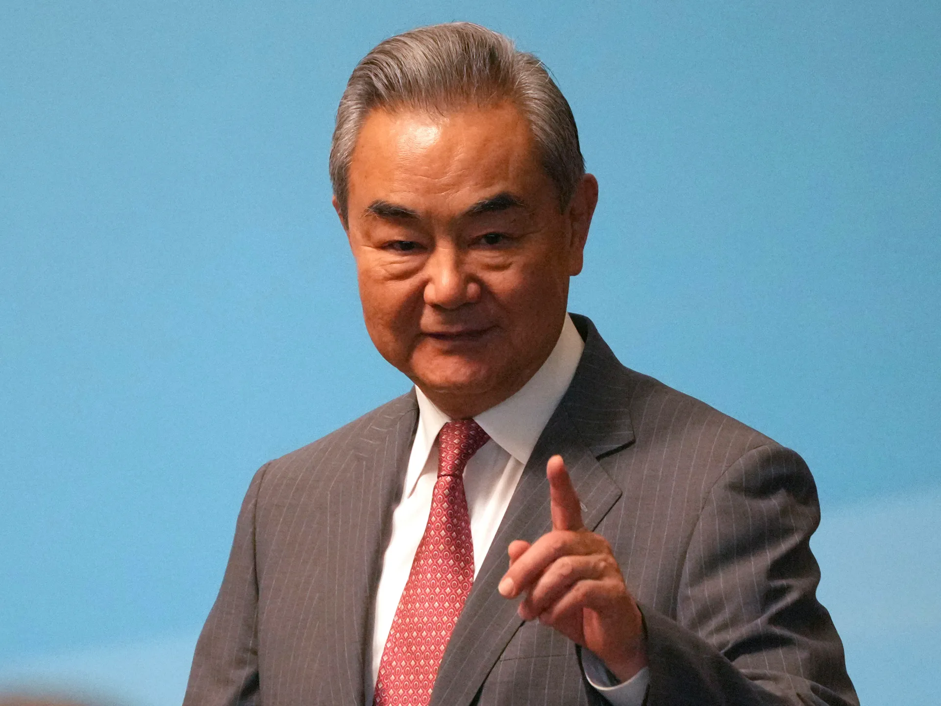 Older man in a gray pinstripe suit and red tie raises one finger against a blue background