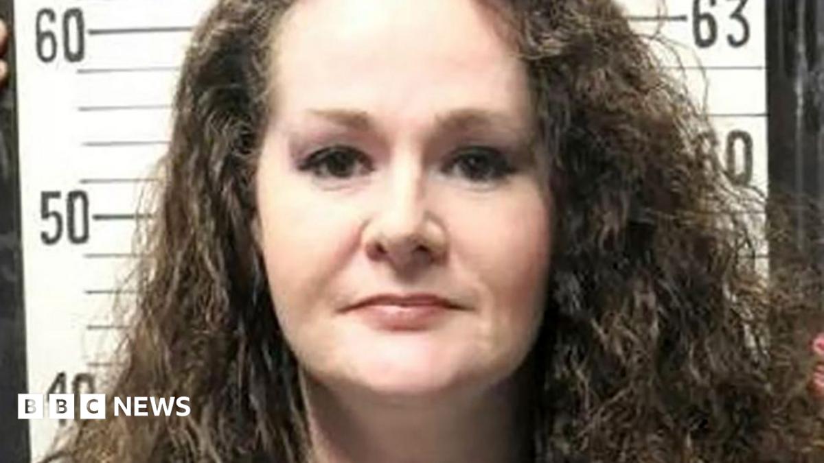 A woman with curly dark hair faces the camera in front of a height chart.