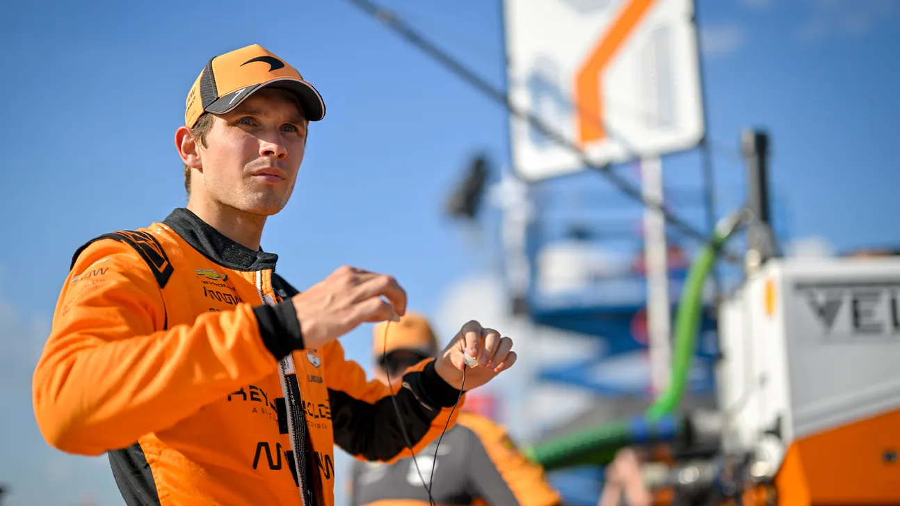 A man in an orange racing suit and cap holds a small wired device outdoors.