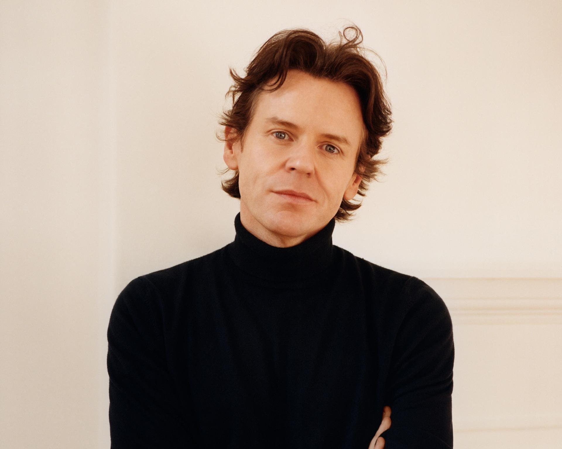 Man with tousled brown hair wearing a black turtleneck against a pale wall