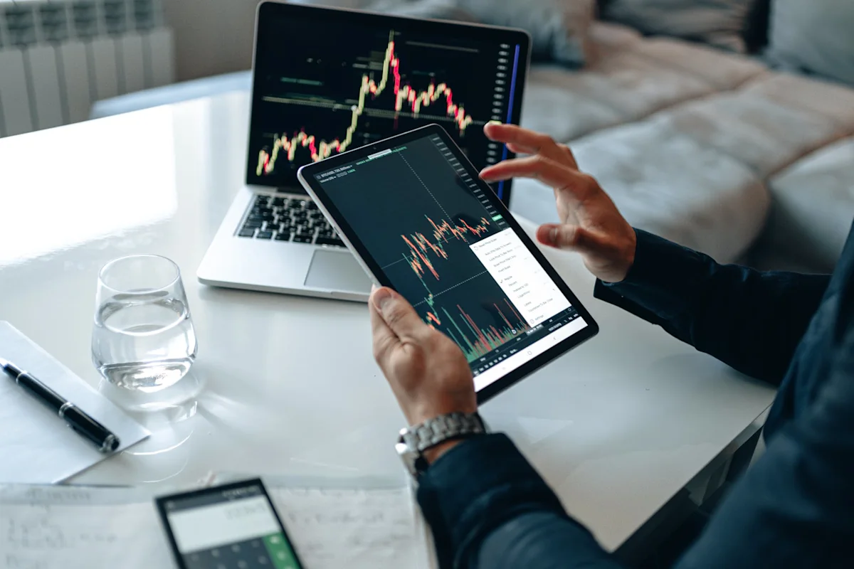 Person reviews stock charts on a tablet beside a laptop at a desk