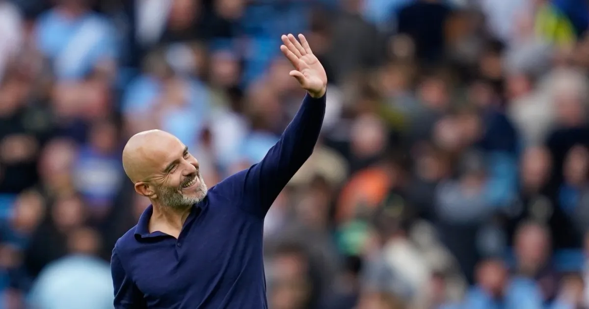 Smiling bald football manager in navy shirt waves to a blurred stadium crowd