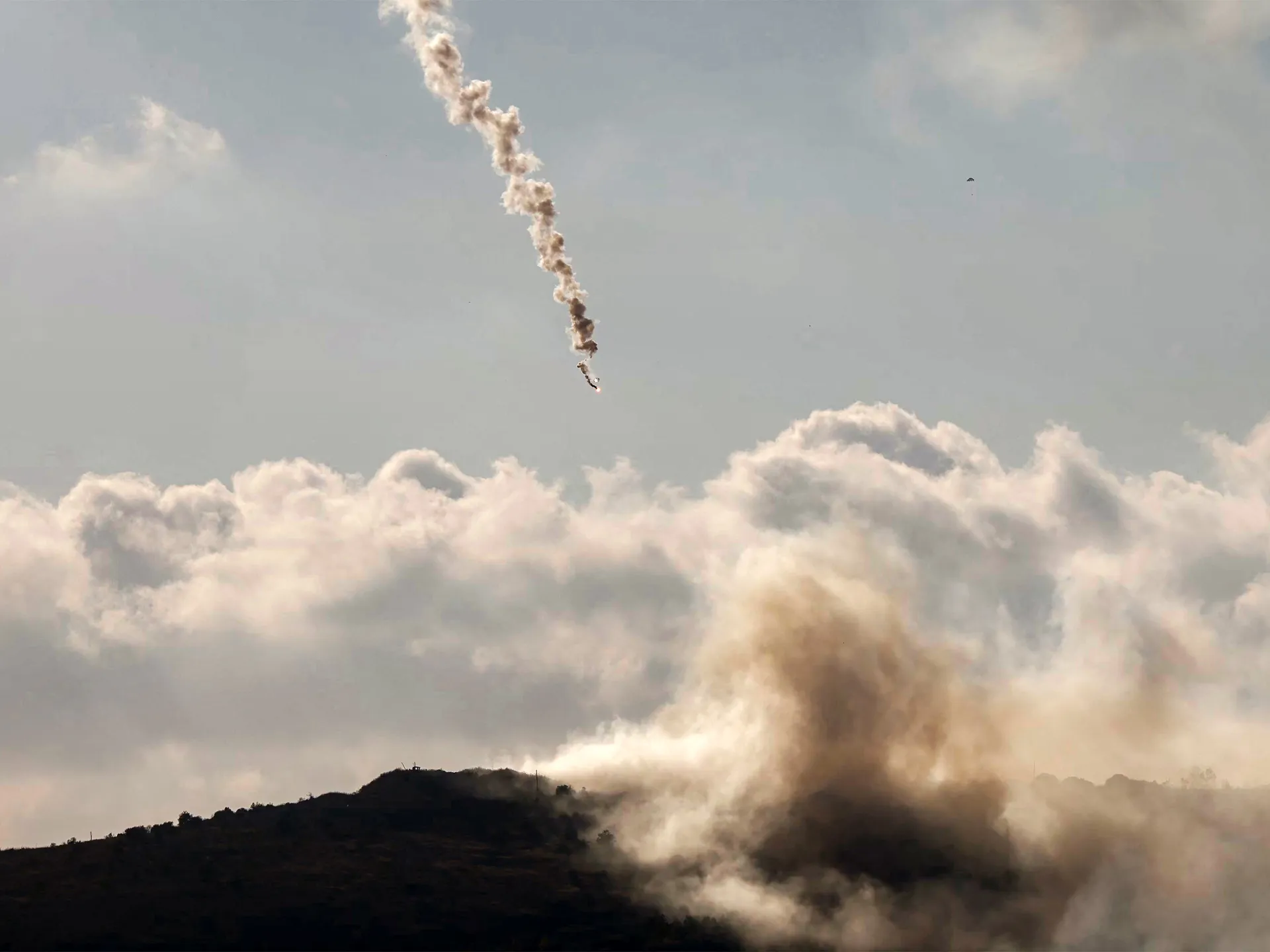 Projectile trails smoke above a dark ridge as dust and clouds billow across the sky