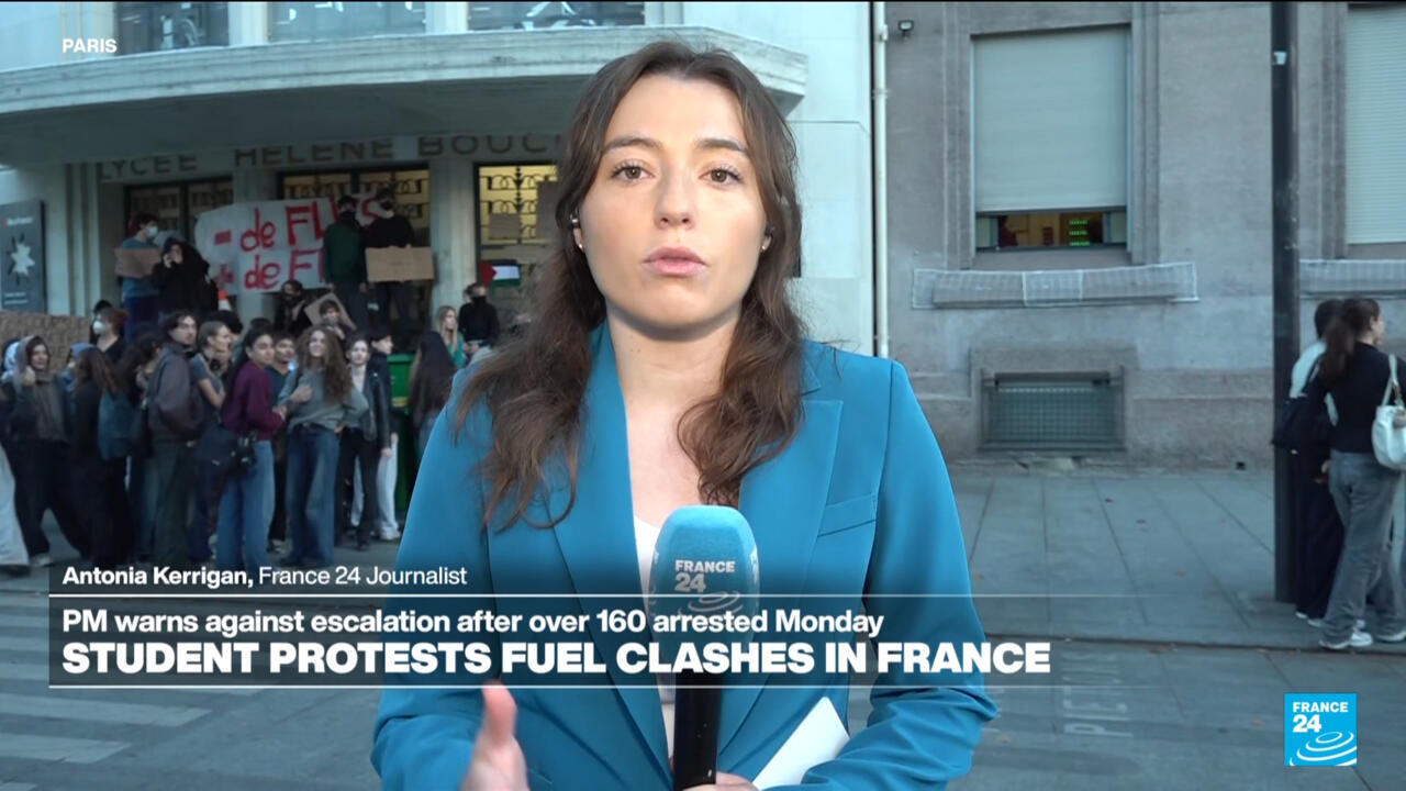 A reporter in a blue blazer speaks outside a school as students gather behind her.