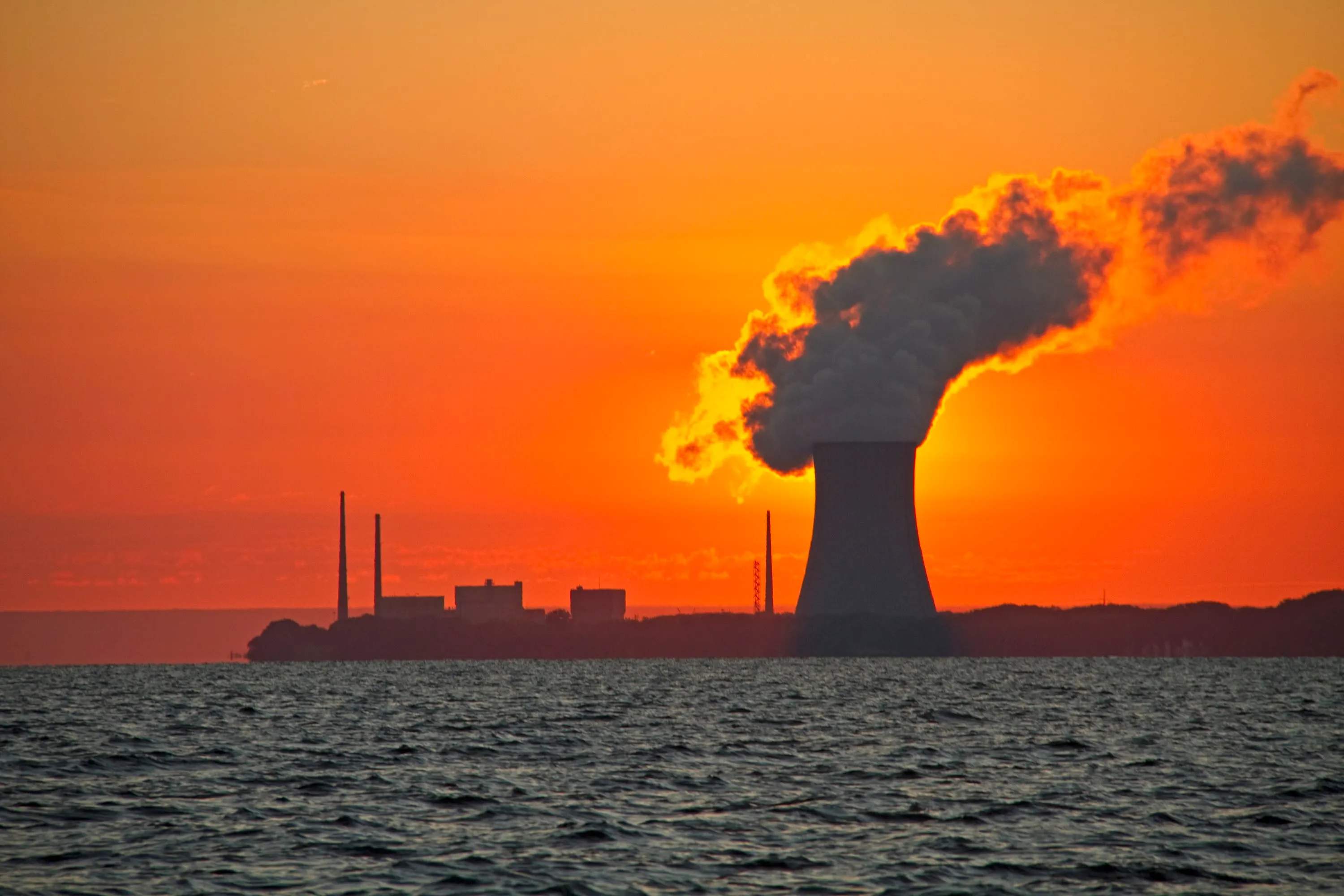 Nuclear power plant silhouetted against a vivid orange sunset, with smoke rising from its cooling tower.
