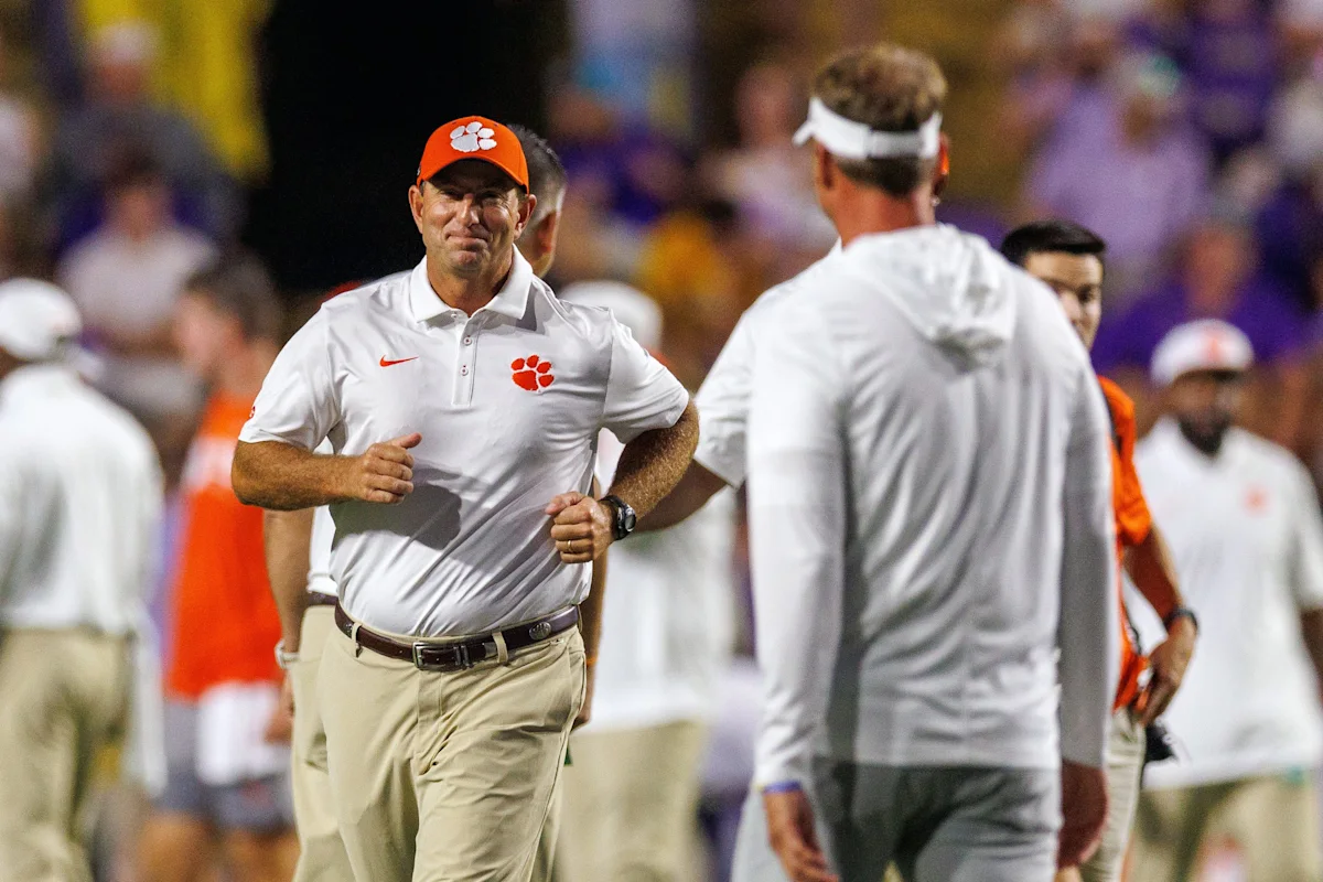 Clemson coach in orange cap jogs past players on a football field during a game