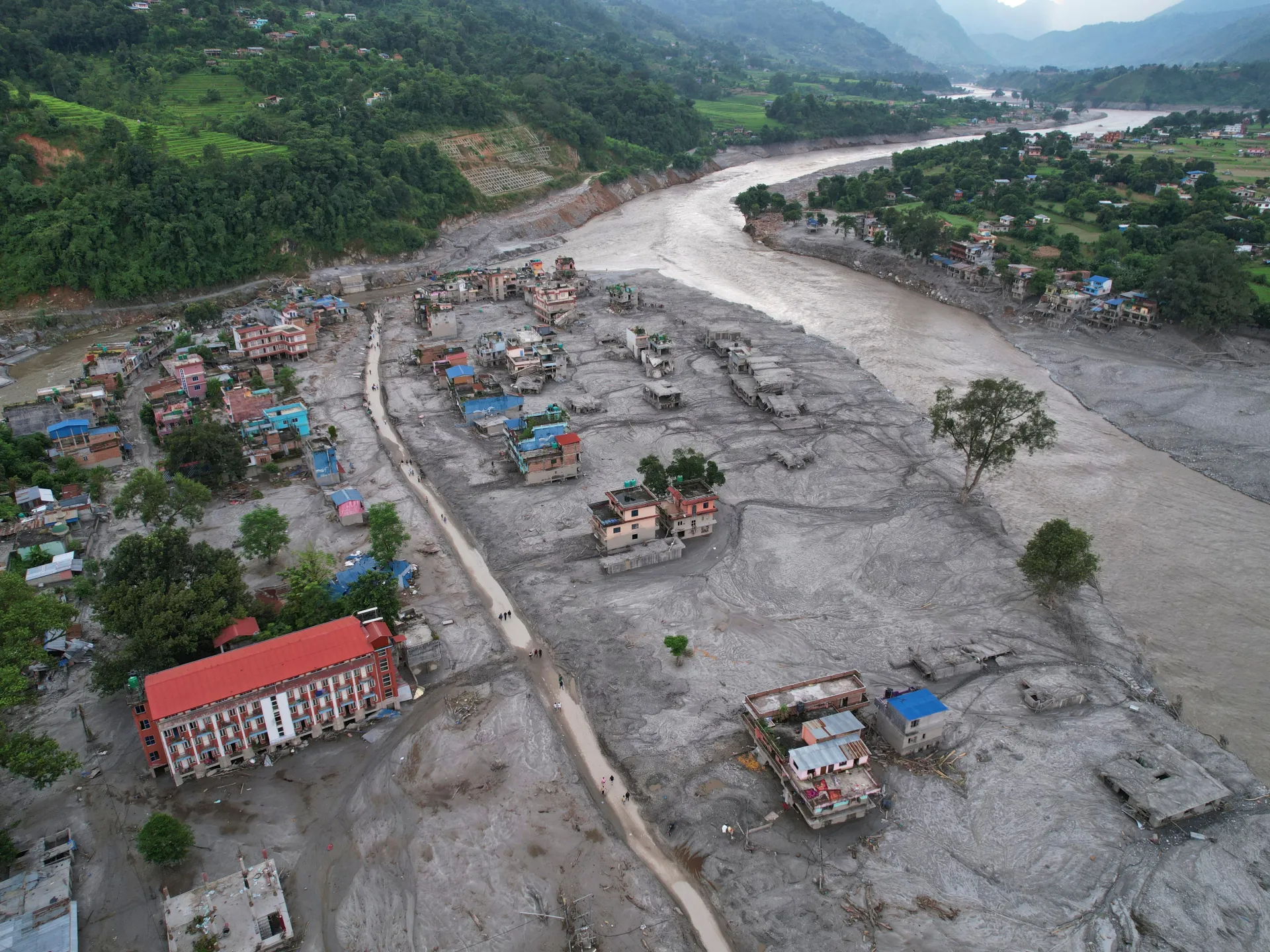Aerial view of a flooded settlement covered in gray sediment beside a swollen river and green hills