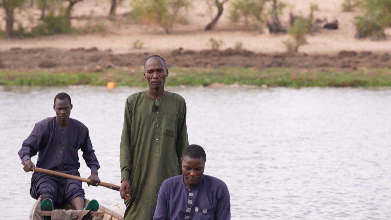 Three men sit and stand in a wooden canoe on a lake, with one holding a paddle.