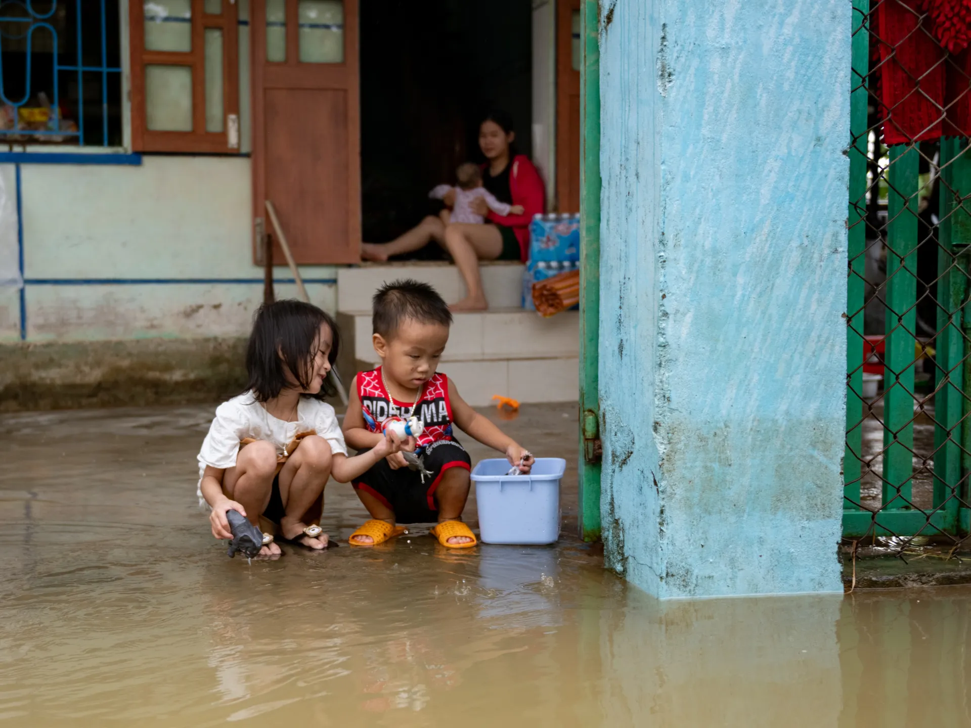 Children sit in floodwater outside a home as an adult holds a baby in the doorway.